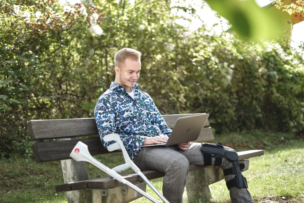 verletzter Patient sitzt glücklich mit Krücken und Laptop auf Schoß im Park